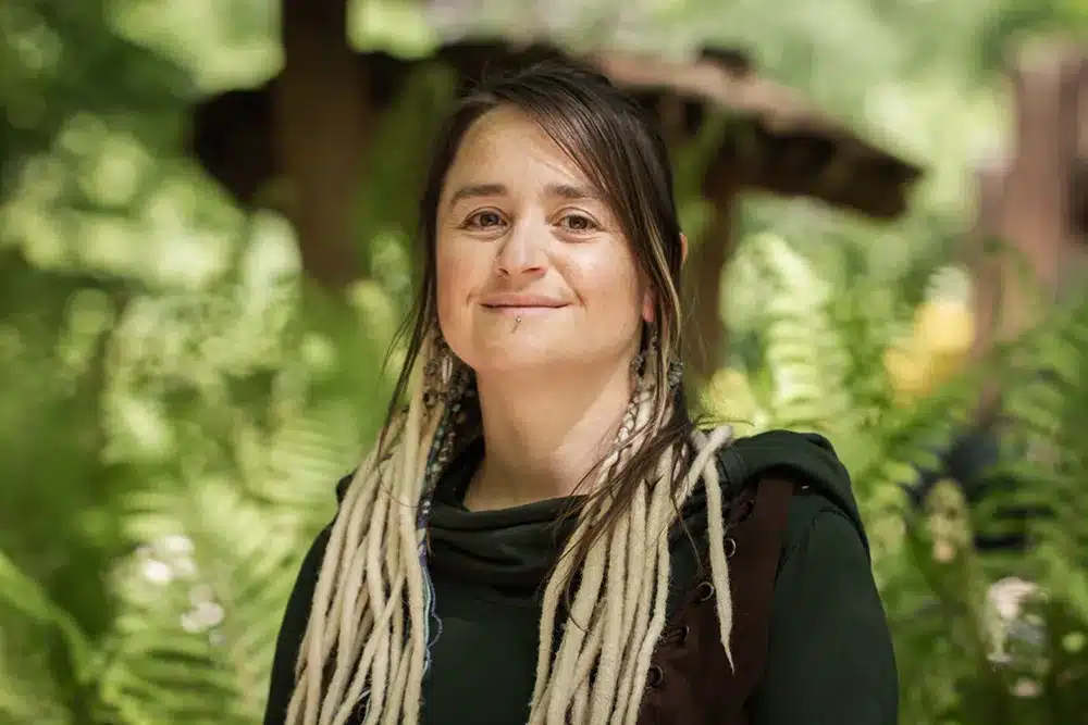 Portrait de Nathalie Poncelet Portrait de Nathalie Poncelet. Jeune femme brune avec des dreads de laine blanc cassé et souriante. Elle est dans un décors de nature avec un champignon sculpté en bois derrière elle.